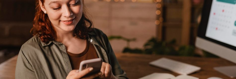 Jovem pesquisando no celular em frente ao computador em casa