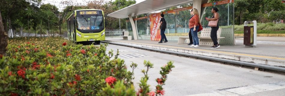 Ônibus se aproxima da estação Lagoa de Piratininga, em Niterói, onde passageiros aguardam sob a cobertura, com canteiro florido em primeiro plano